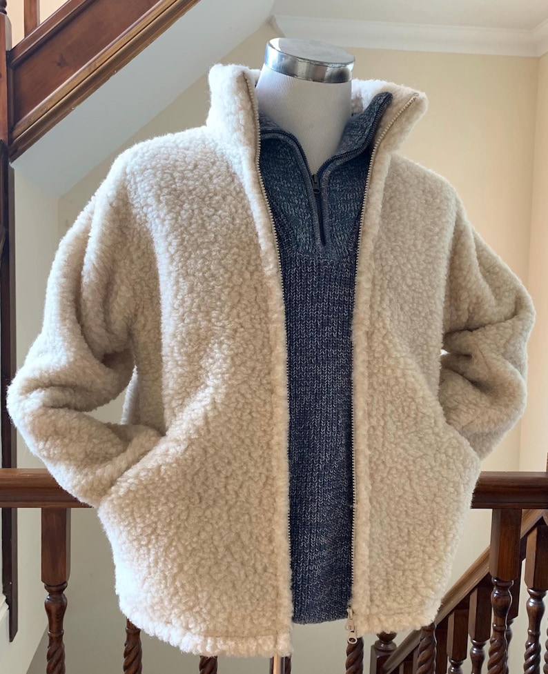 Oatmeal beige unisex wool jacket displayed on a mannequin, featuring a soft textured fabric, full zip front, high collar, and side pockets in an indoor setting