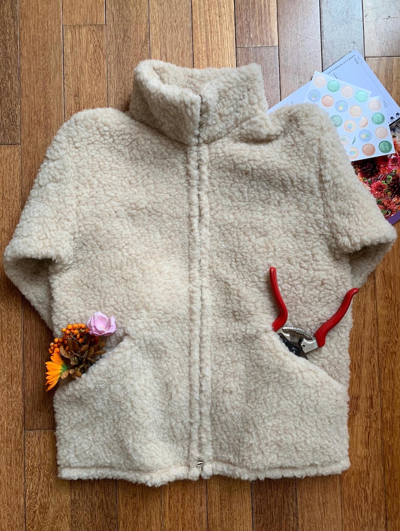 May include: Cream-colored, short-sleeved fleece jacket with a high collar and a full zipper. The jacket has two front pockets, one containing a small bouquet of flowers and the other a pair of red-handled pruning shears.
