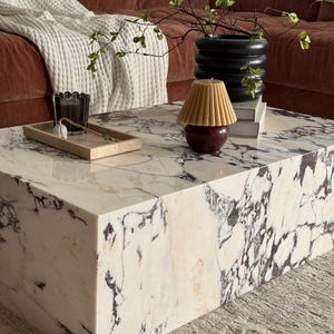 May include: A rectangular marble coffee table with a white and dark purple veined pattern. A small tray with decorative items sits on the table, along with a candle and a black vase with greenery. A brown sofa and white blanket are in the background.