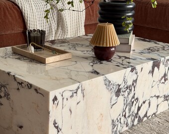 May include: A marble coffee table with a white and dark gray veined pattern. A small tray with decorative items sits on the table, along with a candle and a black vase with greenery. A brown sofa and white blanket are in the background.