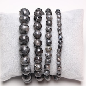 Pode incluir: Cinco pulseiras de contas pretas com tamanhos de contas variados, exibidas em um almofada de veludo cinza.