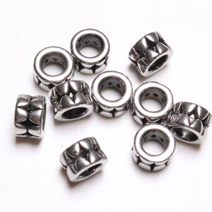 May include: A collection of silver-colored spacer beads. Each bead is cylindrical with a repeating pattern of stylized shapes around the circumference. The beads are scattered on a white surface.