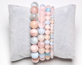 May include: Several beaded bracelets in shades of pink, peach, and blue are displayed on a gray pillow. The bracelets feature round gemstone beads of varying sizes, creating a layered effect. The beads appear to be made of quartz or similar materials.