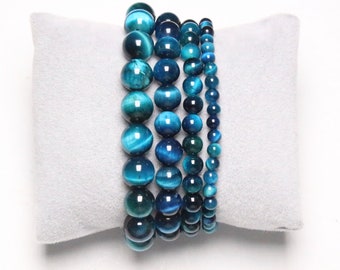 May include: Five blue tiger's eye gemstone bracelets stacked on a gray velvet cushion. The bracelets are made of round beads of varying sizes.