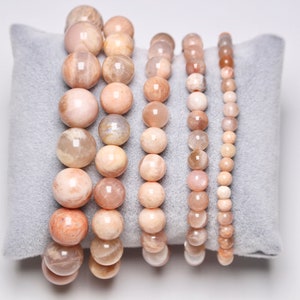 Peut inclure: Un ensemble de six bracelets en pierre de soleil de différentes tailles, avec des perles rondes dans des tons de pêche, blanc et brun clair. Les bracelets sont présentés sur un coussin en velours gris.