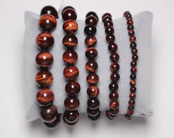 May include: Six red tiger's eye beaded bracelets of varying sizes, displayed on a gray velvet cushion.