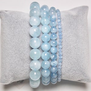 May include: Four aquamarine beaded bracelets stacked on a grey velvet cushion. The bracelets are made of different sized beads, with the largest beads being at the top of the stack.