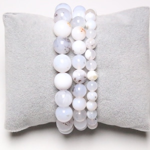Peut inclure: Quatre bracelets en agate blanche en perles empilés sur un coussin de velours gris. Les perles sont rondes et présentent un motif marbré blanc et gris subtil.
