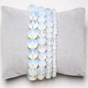 Peut inclure: Cinq bracelets en perles d'opale blanche empilés sur un coussin de velours gris. Les bracelets sont faits de perles rondes et lisses.