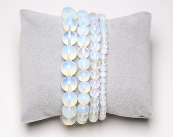 May include: Five white opal beaded bracelets stacked on a gray velvet cushion. The bracelets are made of round, smooth beads.
