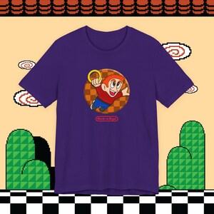 May include: A purple t-shirt featuring a cartoon character with red hair and a red shirt, holding a gold ring. The character is set against a brown and orange checkered background. The text "Retro Ryu" is below the character.