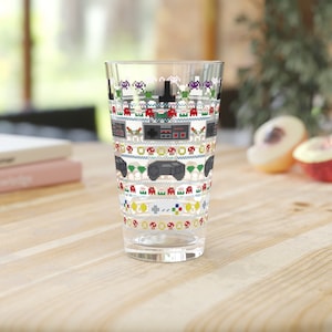 May include: A clear glass with a pixelated design of video game controllers, characters, and game consoles. The design is in a variety of colors, including red, green, blue, yellow, and black.