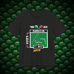 以下が含まれることがあります： レトロなビデオゲームのデザインが施されたダークグレーのTシャツ。スミスという名前のフットボール選手が40番と22番を着ています。デザインには、ピクセル化されたフットボールフィールドとプレイダイアグラム、ピンクのボックスに「PASS」というテキストが含まれています。