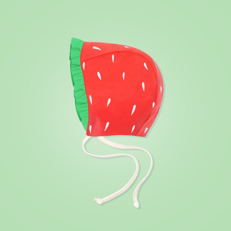 Peut inclure: Un bonnet de b&eacute;b&eacute; en forme de fraise rouge avec des feuilles vertes et des graines blanches. Le bonnet a un lien blanc qui peut &ecirc;tre nou&eacute; sous le menton.