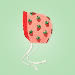 Peut inclure: Un bonnet de b&eacute;b&eacute; rose avec un lien blanc et un imprim&eacute; de fraises. Les fraises sont rouges avec des feuilles vertes.