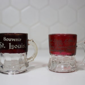 Op de afbeelding: Twee kleine glazen souvenirbekers met handvatten. De beker links is rood met witte letters die zeggen "Souvenir St. Louis." De beker rechts is rood met witte letters die zeggen "Missouri State Election 1976 Voting Souvenir  Jefferson City, Missouri."