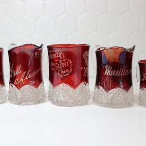 May include: Five red glass souvenir cups with gold lettering. The cups are decorated with a clear glass pattern. The cups are labeled "Sister 1904", "West Alezan", "Mother San George 1904", "Merrillan", and "Revere Beach 1905".