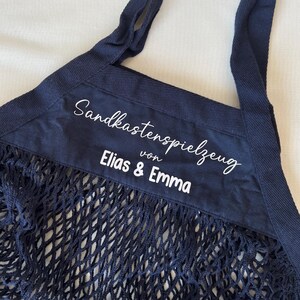 Könnte beinhalten: Eine marineblaue Netztasche mit verstärktem Oberteil. Der obere Bereich zeigt weißen Text mit der Aufschrift "Sandkastenspielzeug von Elias & Emma". Die Tasche hat zwei marineblaue Griffe.