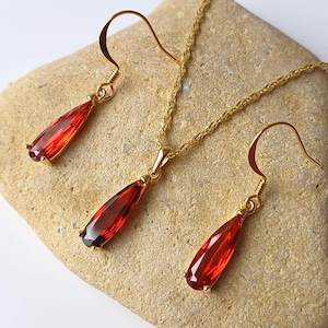 May include: A gold-coloured jewellery set comprising a necklace and earrings. Each item displays a teardrop-shaped, faceted, red gemstone. The necklace has a fine chain, and the earrings have curved hooks.
