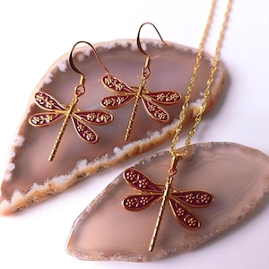 May include: A gold-tone jewellery set with a dragonfly motif. The set comprises a necklace and earrings. The dragonfly wings are a deep red with gold floral detailing. The jewellery is presented on a light pink and brown surface.