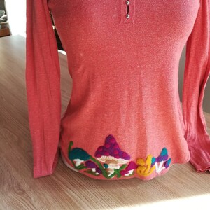 Peut inclure: Un chemisier à manches longues de couleur corail avec un col en V et une rangée de boutons argentés. Le chemisier est orné d'un motif brodé de champignons colorés sur l'ourlet inférieur.