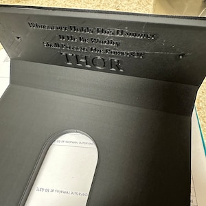 Peut inclure: Un présentoir noir imprimé en 3D avec le texte "Whosoever Holds This Hammer If He Be Worthy Shall Possess The Power Of THOR". Le présentoir a une découpe incurvée et est conçu pour contenir un objet.