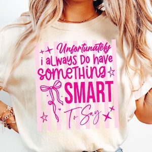 May include: A cream-colored t-shirt featuring a graphic with pink vertical stripes and the text "Unfortunately i ALWAYS Do have SOMETHING SMART To Say" in pink lettering, accompanied by a pink ribbon bow illustration and small star accents.