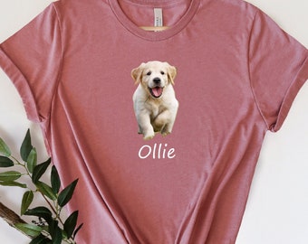 May include: A pink t-shirt with a white graphic of a golden retriever puppy. The puppy is looking at the camera and has its mouth open. The text "Ollie" is printed below the puppy.