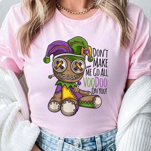 May include: A pink t-shirt with a cartoon voodoo doll wearing a purple and green jester hat. The doll has a stitched-up face and is holding a needle and thread. The text on the shirt reads "Don't Make Me Go All Voodoo On You!"