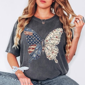 May include: Dark gray t-shirt featuring a large butterfly design. One wing displays the American flag, while the other showcases a floral pattern. The shirt has a relaxed fit and short sleeves.