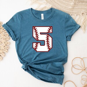 May include: A teal blue t-shirt with a white baseball number "5" with red stitching. The number is centered on the shirt.