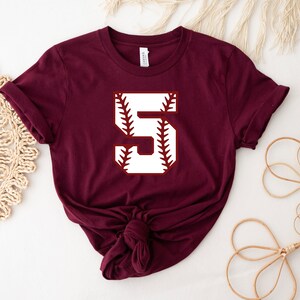 May include: Burgundy short-sleeved t-shirt with a white baseball-themed number "5" graphic.