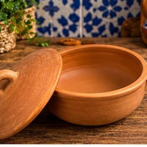 May include: A round, terracotta-colored clay cooking pot with a matching lid. The pot features two small side handles, while the lid has a central loop handle. The surface of the clay is smooth with visible circular textures from the crafting process.