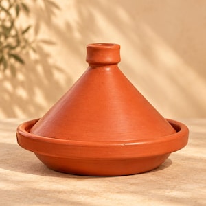 May include: A terracotta tagine cooking vessel featuring a wide, circular base and a tall, conical lid with a small knob at the top. The surface of the clay pot is smooth and uniform in a warm, earthy reddish-brown colour, showcasing a traditional design.