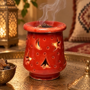 May include: A red ceramic incense burner with a glossy finish, featuring cut-out shapes of stars, moons, and trees. The cylindrical vessel has a wide top rim and emits a thin trail of smoke from the burning resin placed inside the center bowl.