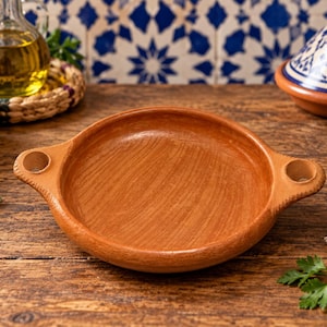 Puede incluir: Un plato de barro de color terracota con un interior liso y poco profundo, y dos pequeñas asas circulares en los lados opuestos. El plato presenta un acabado natural y terroso con una textura visible y un borde ancho y abierto.
