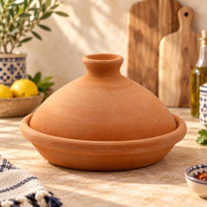Puede incluir: Un recipiente de cocina tipo tajine de terracota con una base circular ancha y una tapa cónica con un pequeño mango redondeado en la parte superior. La superficie de la olla de barro es mate, sin esmaltar y tiene un color rojizo natural.