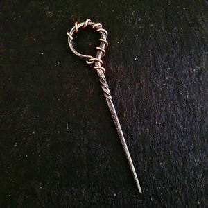 Puede incluir: Un palillo para el pelo de metal retorcido con un bucle de alambre de cobre en la parte superior. El palillo es de color plateado y el bucle es de color cobre cálido.