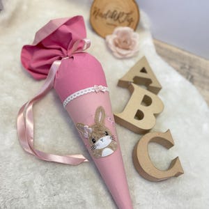 Könnte beinhalten: Eine rosa Schultüte aus Stoff mit einem Hasen-Applikation. Der Kegel ist mit einer weißen Spitzentrimmung und einem rosa Band verziert. Die Oberseite des Kegels ist gerafft und mit einem rosa Band gebunden. Holzbuchstaben ABC im Hintergrund.