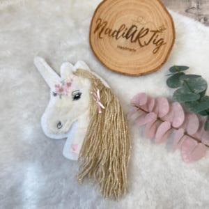Könnte beinhalten: Ein weißer Einhornkopf mit goldener Mähne, rosa Blumenkranz und kleiner rosa Schleife. Das Einhorn hat ein Herz auf der Wange. Im Hintergrund befindet sich eine Holzscheibe mit dem Text "NadiARTig Handmade".