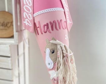 Könnte beinhalten: Rosa Schultüte mit einem weißen Band, auf dem "Schulkind" und das Jahr "2023" stehen. Die Tüte hat einen rosa-weißen Besatz und den Namen "Hanna" bestickt. Ein brauner und weißer Pferdekopf mit einer langen weißen Mähne ist an der Tüte befestigt.