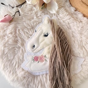 Könnte beinhalten: Ein weißer Einhornkopf aus Filz mit einer Mähne aus beigefarbenem Garn und einem Blumendesign. Das Einhorn hat schwarze Wimpern und ein detailliertes Auge. Der Hintergrund ist ein flauschiges, cremefarbenes Material mit einer rosa Blume und einem Holzstück im Hintergrund.