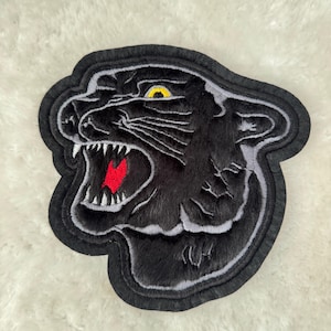 Könnte beinhalten: Schwarzer gestickter Pantherkopf-Patch mit rotem Mund und gelbem Auge. Der Patch befindet sich auf einer weißen, flauschigen Oberfläche.