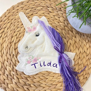 Könnte beinhalten: Weißer Einhornkopf mit einer lila Mähne und einem lila Schwanz, bestickt mit rosa Blumen und dem Namen "Tilda" auf einem weißen Banner.