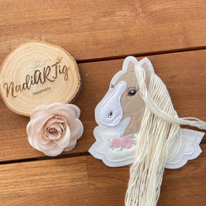 Könnte beinhalten: Eine beige Filz-Pferdekopf-Applikation mit einer weißen Filz-Mähne und gestickten Blumendetails. Der Pferdekopf ist auf einem weißen Filz-Grund mit einer langen weißen Filz-Mähne befestigt.