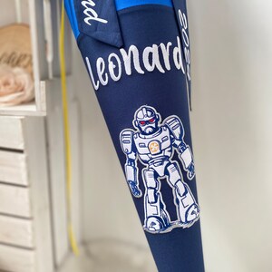 Könnte beinhalten: Ein blauer und marineblauer Schultüte mit einer weißen Robotergrafik und dem Text "Leonard 2023" und "Schulkind" bestickt.