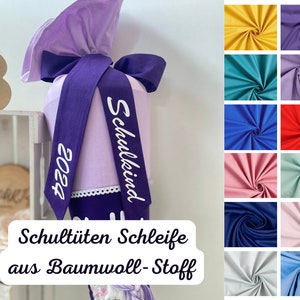 Könnte beinhalten: Ein lilafarbener Stoffschultüte mit weißem Text, der "Schulkind" und "2023" lautet. Die Tüte ist mit einem lilafarbenen Band und weißer Spitze verziert. Der Text "Schultüten Schleife aus Baumwolle-Stoff" ist unter der Tüte abgebildet.