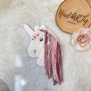 Könnte beinhalten: Ein weißer Einhornkopf mit Horn und rosa und lila Garnhaar. Das Einhorn hat einen rosa Blumenkranz und ein Herz auf der Wange. Das Bild enthält auch eine Holzscheibe mit dem Text "NadiikTig Handmade" und eine rosa Blume.