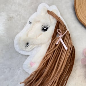 Könnte beinhalten: Weißer Plüschpferdekopf mit braunem Garnhaar, einer rosa Schleife und gestickten Details. Das Pferd hat ein weiches, strukturiertes Aussehen mit einer hellrosa Nase und schwarzen Augen. Dieser Artikel ist wahrscheinlich ein Dekorationsstück oder ein Kinderspielzeug.