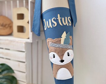 May include: A blue fabric cone with a fox applique wearing a headdress. The cone has the name "Justus" embroidered on it in white.